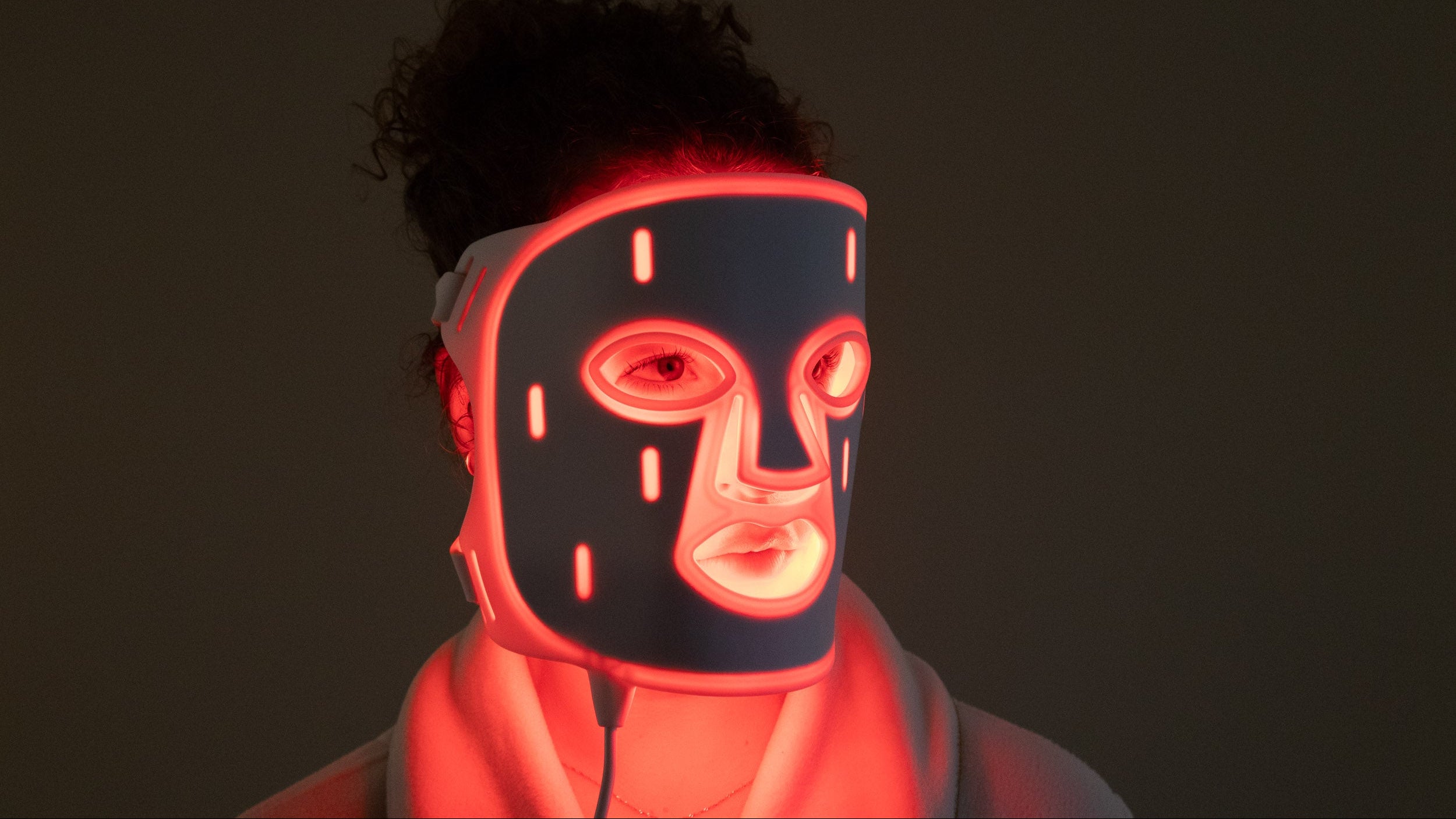 Close-up front view of woman wearing PHOS LED face mask with red light therapy glowing against dark background